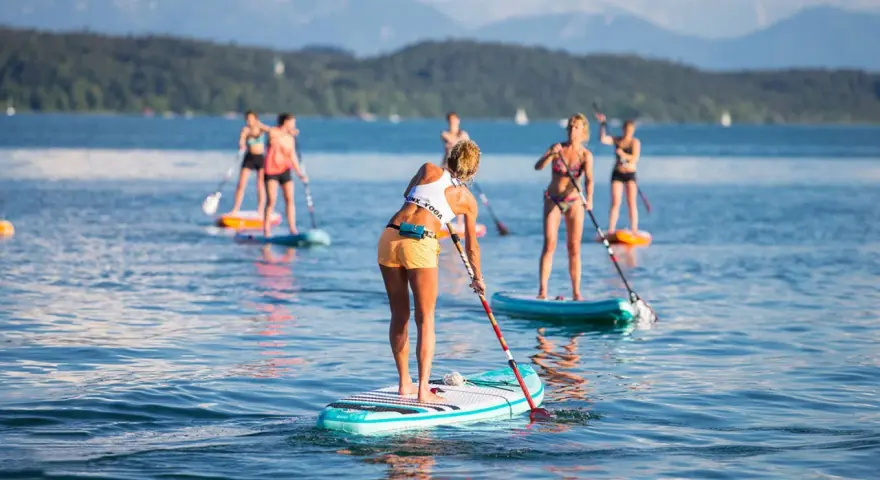 Eine Gruppe von Frauen genießt das Stand-Up-Paddling auf einem See. Sie stehen auf ihren Boards und paddeln durch das ruhige Wasser, während im Hintergrund Berge und Bäume zu sehen sind.