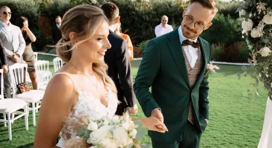 Para Młoda trzyma bukiet, a Pan Młody w zielonym garniturze uśmiecha się do niej. To piękny moment podczas urlopu okolicznościowego ślub.
