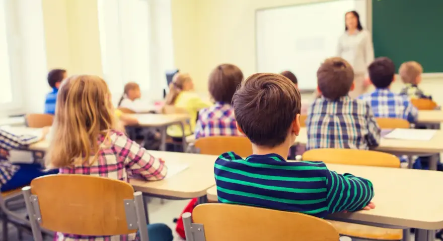 Dzieci siedzą w ławkach podczas lekcji religii w szkole, słuchając nauczycielki stojącej przy tablicy.