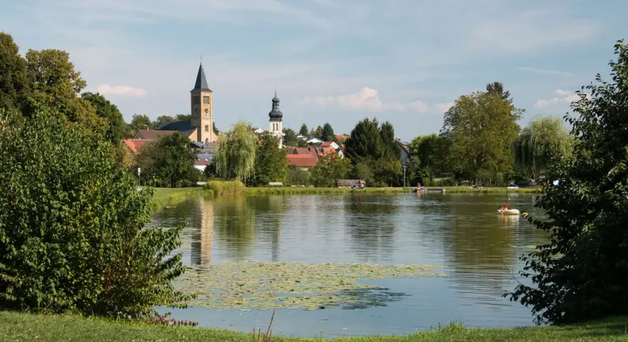Idyllischer Elsenzer See mit Seerosen, Bäumen und einer Kirche im Hintergrund. Ein Boot mit zwei Personen treibt auf dem Wasser.