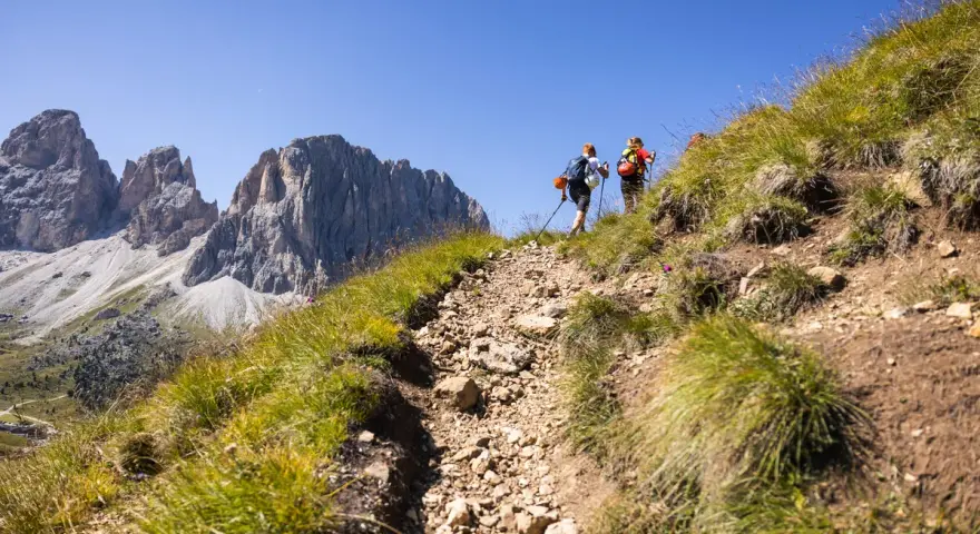 Due escursionisti affrontano un sentiero roccioso sui **cammini in Trentino**, con imponenti vette sullo sfondo.