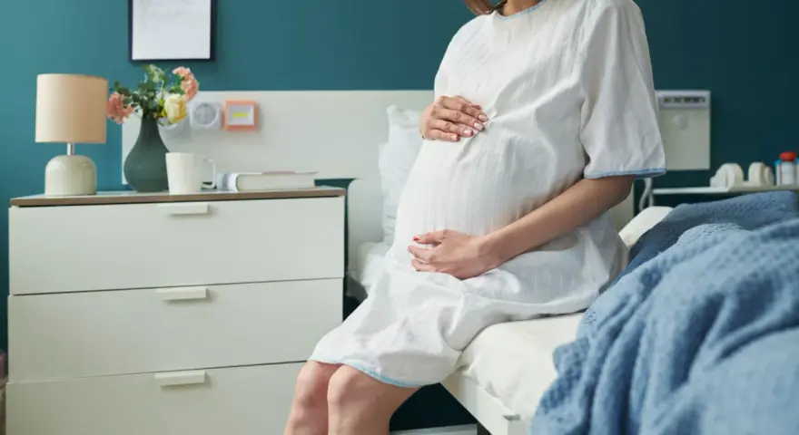 Kobieta w szpitalnej koszuli, z dłońmi na brzuchu, czeka na poród w 3 trymestrze.