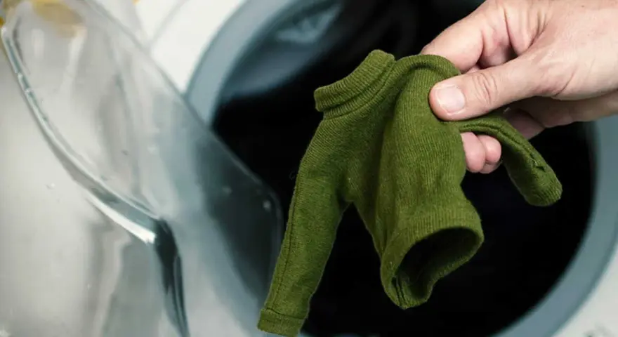 Main verte, petite, tenue dans une main, devant une machine à laver ouverte. Le coton rétrécit il au lavage ?