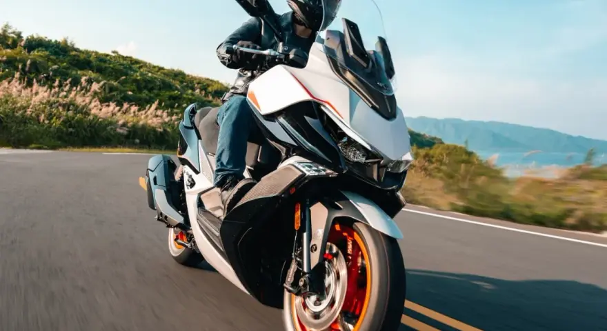 Kymco Xciting 400i dane techniczne: skuter z kierowcą na krętej drodze, z widokiem na góry i jezioro w tle.