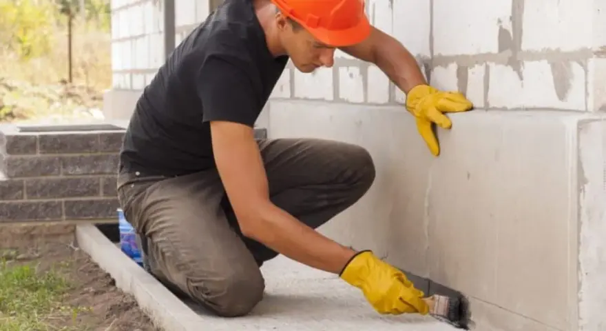 Obrero con casco naranja aplica producto para impermeabilizar pared exterior de bloque de hormigón.