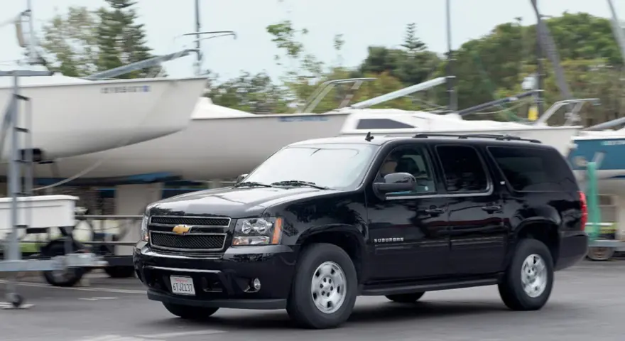 Czarny Chevrolet Suburban jedzie obok przyczep z łodziami, w tle drzewa.