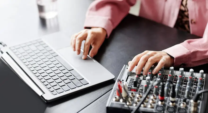Osoba z tatuażami na dłoniach reguluje suwaki na mikserze, obok laptopa. Dowiedz się, jak podłączyć mikser do laptopa.