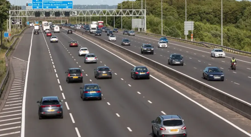 Autostrada z ruchem lewostronnym, pełna samochodów i ciężarówek. Znaki drogowe wskazują M1, A405 i M25.