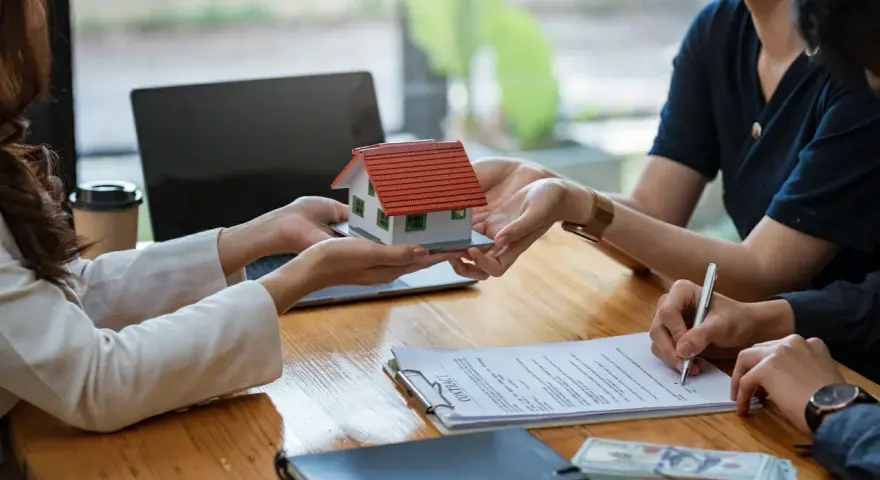 Une maison miniature est remise, un contrat est signé, et de l'argent est posé sur la table, suggérant une transaction immobilière et un chèque de caution.