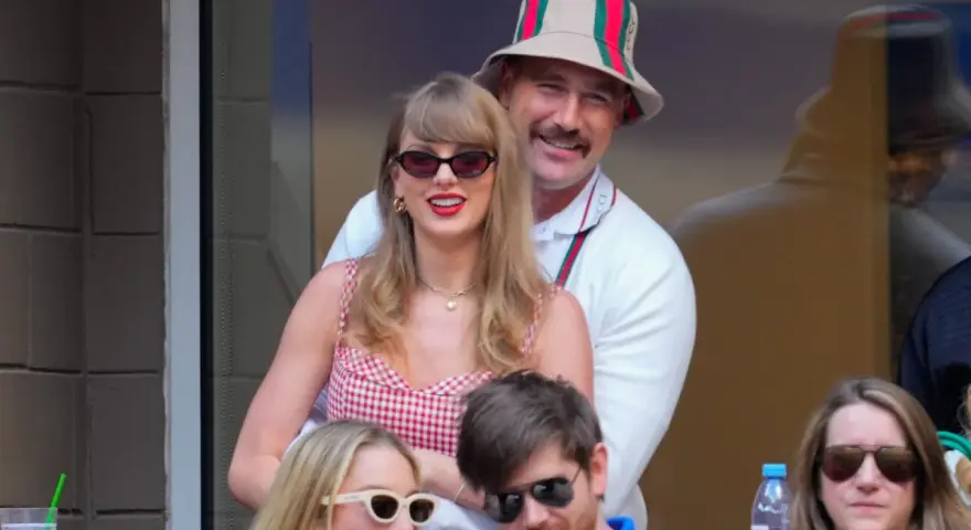 Travis Kelce e Taylor Swift sorridono, lei con un vestito a quadri rossi, lui con un cappello Gucci.
