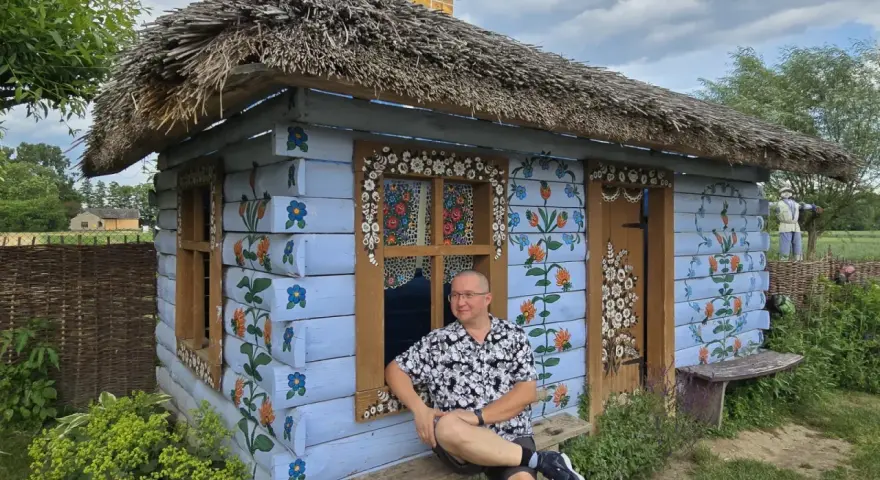 Mężczyzna siedzi na ławce przed malowaną chatką w Zalipiu. Kolorowe kwiaty zdobią ściany, a słomiany dach dodaje uroku. To jedna z wielu zalipiańskich atrakcji.