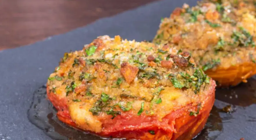 Pomodori gratinati in padella, con croccante panatura alle erbe e formaggio, serviti su una pietra scura.