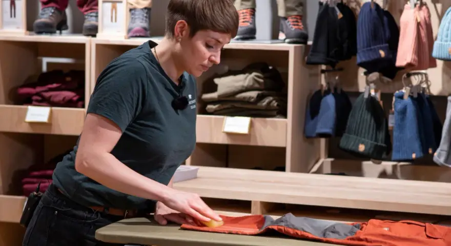 Mitarbeiterin pflegt orangefarbene Hose, damit sie für den Verkauf wie neu wachsen kann. Im Hintergrund sind Kleidung und Mützen ausgestellt.