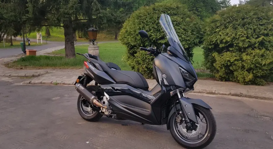 Czarny skuter Yamaha XMAX 300 zaparkowany na tle zieleni i drzew.