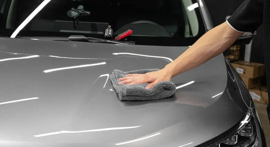 A hand buffs a car's hood with a microfiber cloth, applying a ceramic wax for a deep shine.