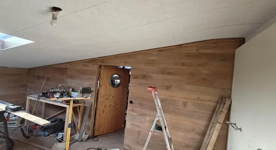 Taket är målat OSB. Rummet är under renovering med träpanel på väggarna, en dörr med runt fönster och byggmaterial utspritt.