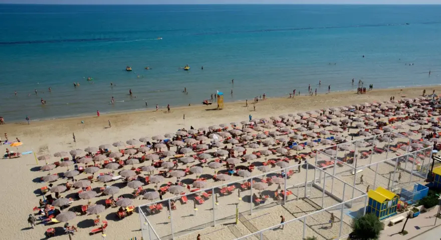 A crowded beach with rows of umbrellas and sunbathers, and people swimming in the blue sea.