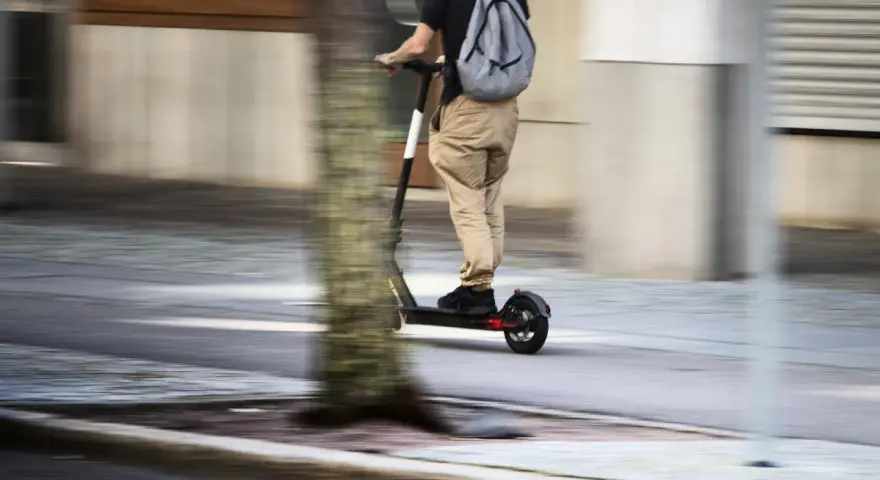 En person på elsparkcykel, med ryggsäck, susar fram på trottoaren. Frågan om får man åka två på elsparkcykel är relevant här.