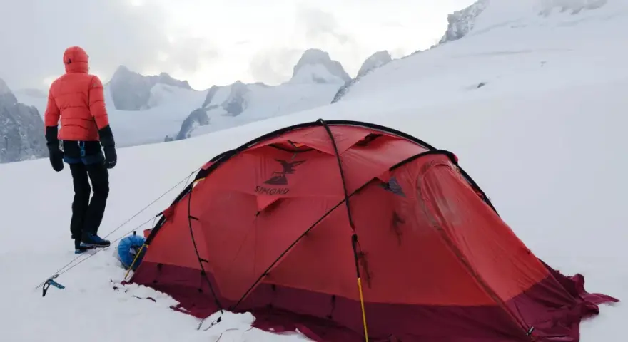 Alpinista w czerwonej kurtce stoi obok namiotu na tle ośnieżonych szczytów. W oddali widać groźne góry, gdzie mogą wystąpić lawiny w Alpach.