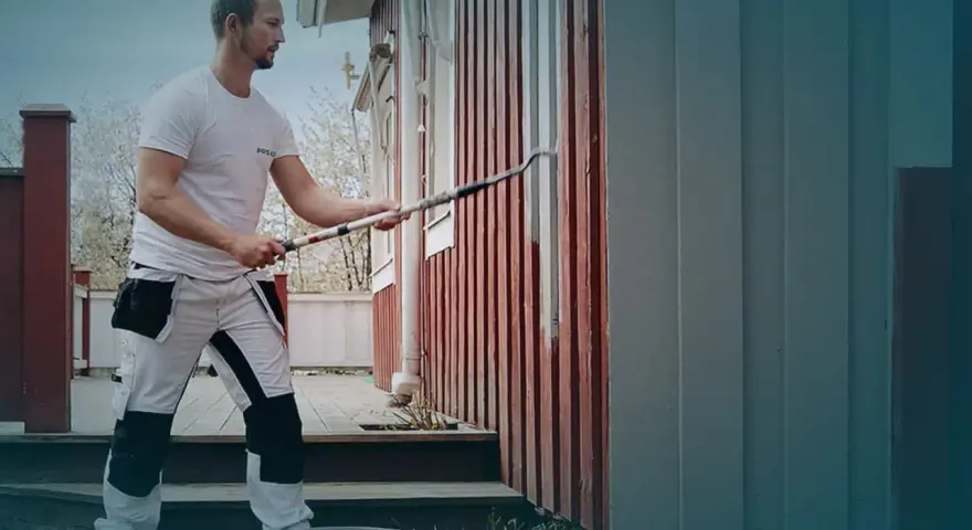 Man i arbetskläder använder en lång roller för att måla huset. En hink med färg står på gräset.