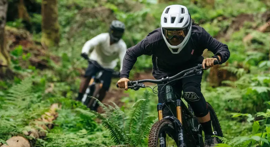 Riders in helmets and protective gear shredding a forest trail. This image shows what to wear mountain biking: full-face helmets, goggles, long sleeves, and knee pads.