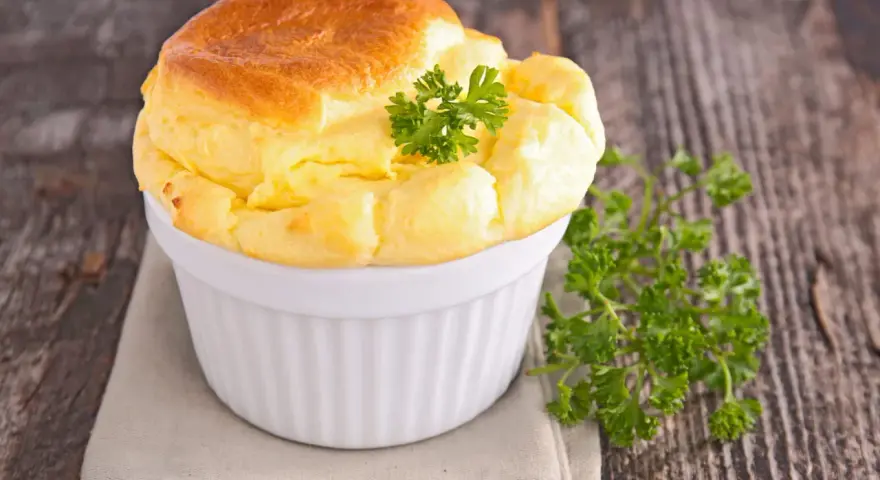 Un soufflé au fromage doré et gonflé, garni de persil frais, dans un ramequin blanc sur une serviette grise.