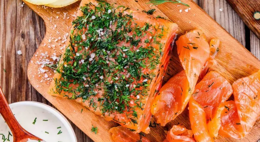 En bit gravad lax, generöst toppad med dill och flingsalt, ligger på en träskärbräda bredvid skivor av lax och en skål med sås.