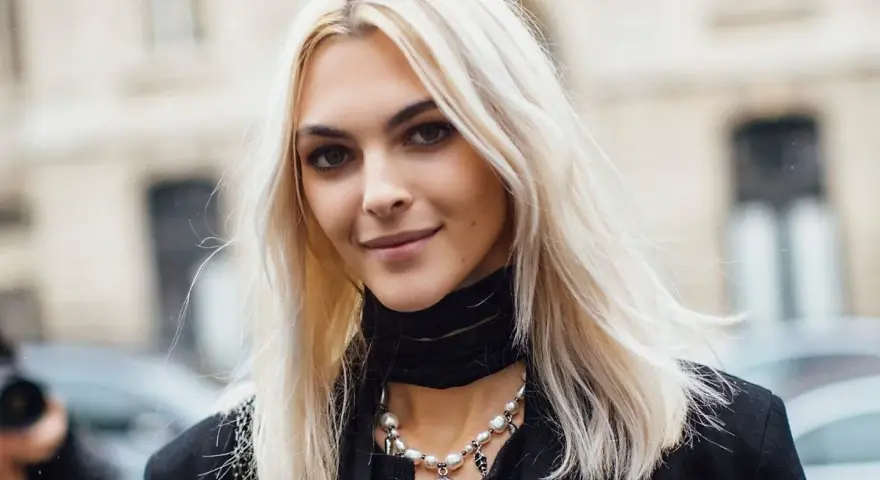 Kobieta z platynowym blondem, w czarnym golfie i naszyjnikiem z pereł, uśmiecha się lekko.