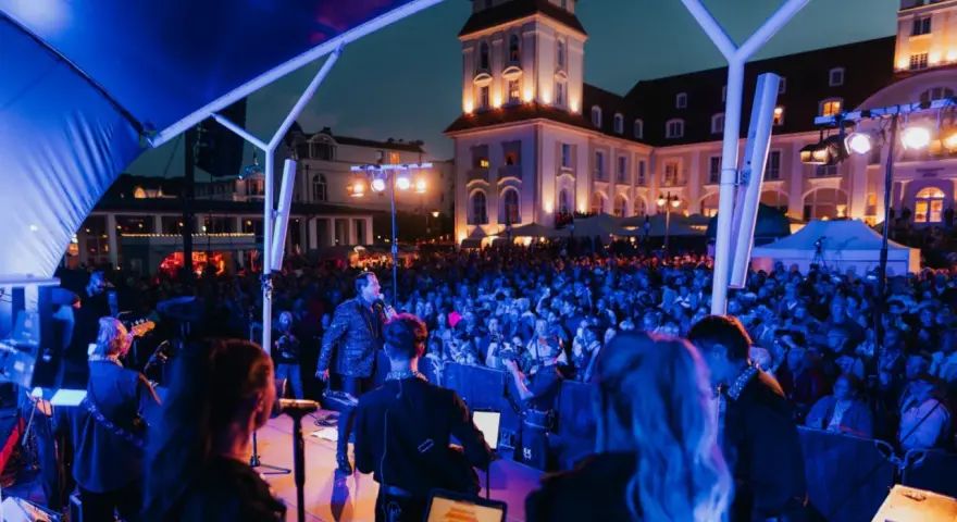 Konzert mit Sänger und Band auf der Bühne, beleuchtet von blauen Lichtern. Im Hintergrund die beleuchtete Architektur und eine Menschenmenge bei Veranstaltungen am Binz Kurplatz.