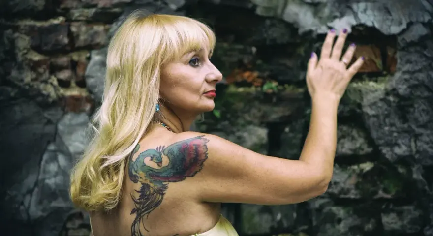 Blondynka z tatuażem feniksa na plecach opiera się o kamienną ścianę. Czyżby moda na tatuaże mijała, a ona wciąż pielęgnuje swoje?