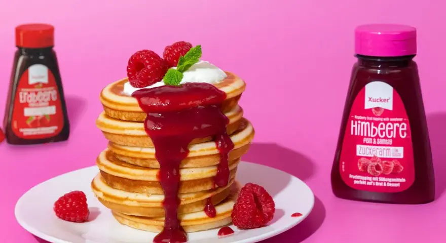 Ein Stapel zuckerfreie pancakes mit Himbeersauce und frischen Beeren. Daneben Flaschen mit Erdbeer- und Himbeer-Topping.