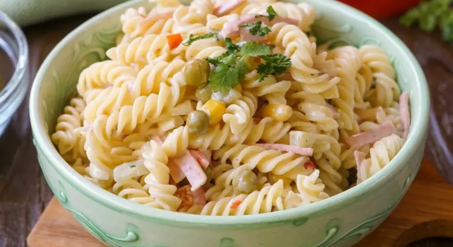 Ein cremiger Nudelsalat mit Erbsen, Mais und Schinkenwürfeln, der an einen herzhaften Fleischsalat erinnert. Garniert mit Petersilie.