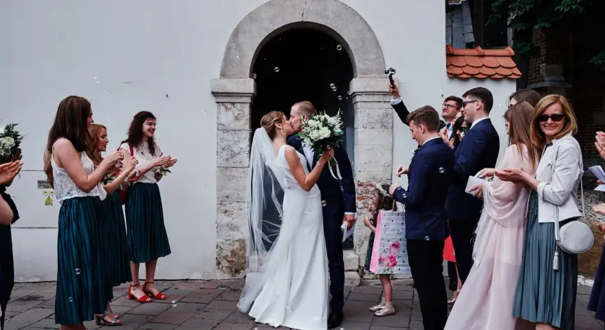 Para młoda całuje się przed kościołem, goście sypią bańki mydlane. To piękny ślub jednostronny, pełen radości i miłości.