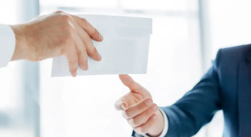 Main d'un homme en costume remettant une enveloppe blanche à une autre main. Peut-être une mise en demeure URSSAF avec un délai de paiement.