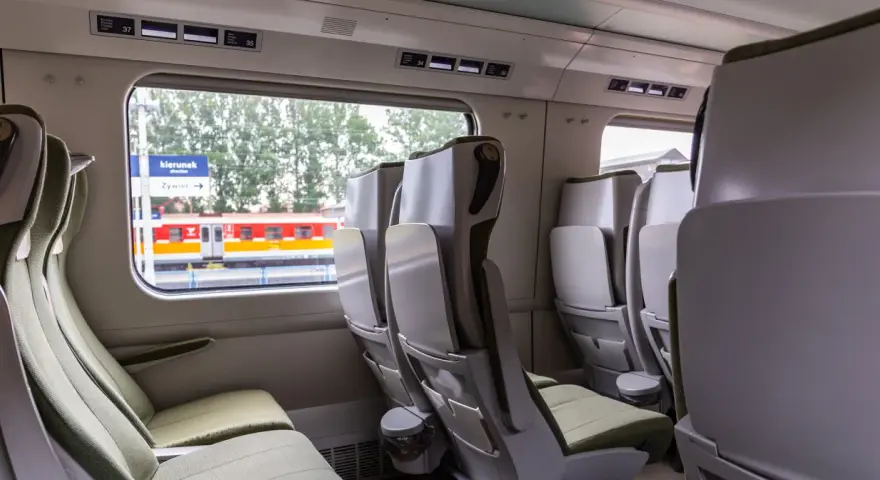 Wnętrze wagonu bez przedziałów IC. Rozkład miejsc z zielonymi siedzeniami i stolikami. Za oknem peron i pociąg.
