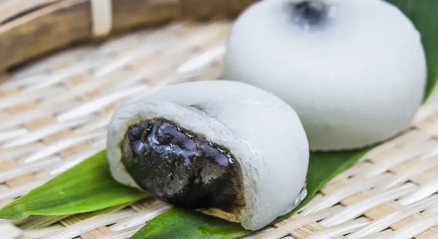 Dwa białe mochi, jedno przekrojone, ukazujące ciemne nadzienie. Podane na bambusowej tacce z liśćmi.