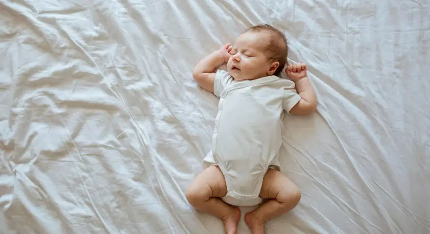 Śpiący, 3 tygodniowy noworodek w białym body leży na pogniecionej białej pościeli.