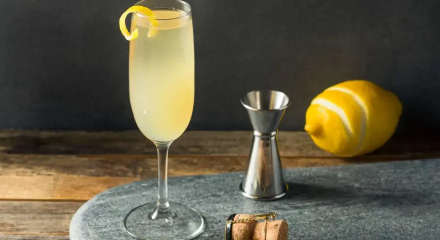 Elegancki drink z szampanem, ozdobiony skórką cytryny, obok kieliszka stoi miarka barmańska, cytryna i korek od szampana.