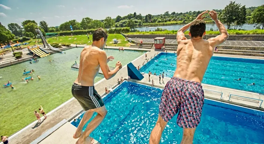 Zwei Jungen springen vom Sprungbrett in den Badesee Bremen. Im Hintergrund sind Wasserrutschen und viele Badegäste zu sehen.