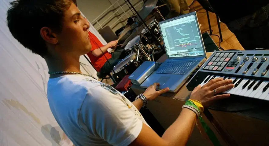 Junger Mann am Laptop und Keyboard, der Musik produziert. Ein Schlagzeuger im Hintergrund. Perfekt für ein Musikproduktion Studium.