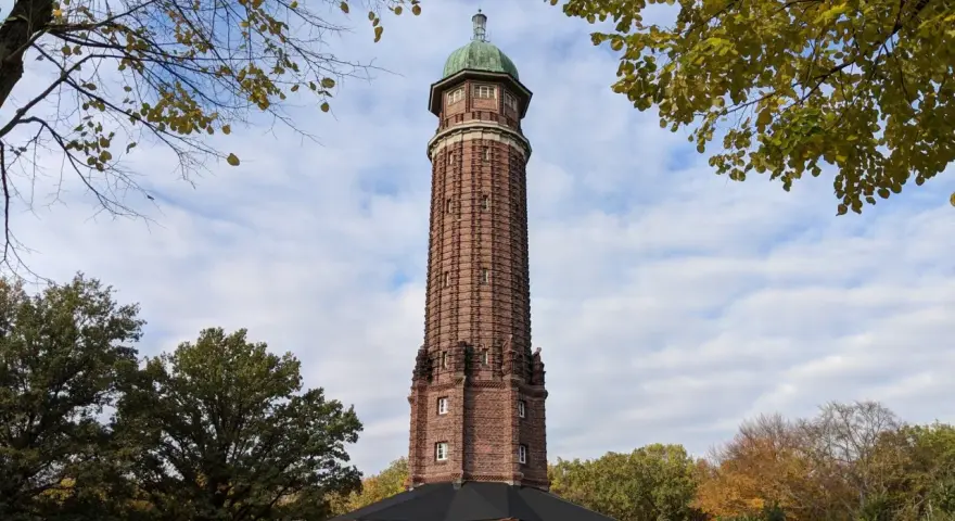 Der Aussichtsturm im Volkspark Jungfernheide ragt mit seinem roten Backstein und grünen Kuppeldach in den Himmel. Herbstlaub umrahmt die Szene.