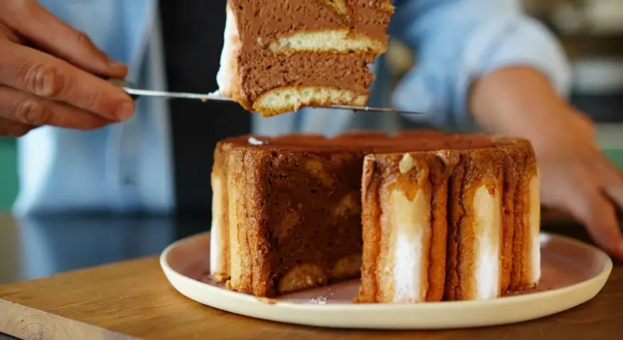Une part de charlotte tiramisu, aérienne et chocolatée, est prélevée d'un gâteau entier.