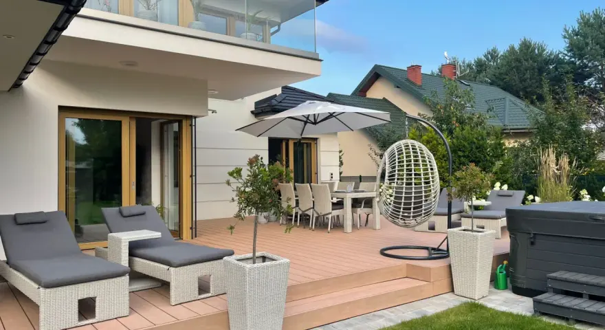 Nowoczesny taras z leżakami, stołem i krzesłami, wiszącym fotelem i jacuzzi. Idealne miejsce na relaks na świeżym powietrzu.