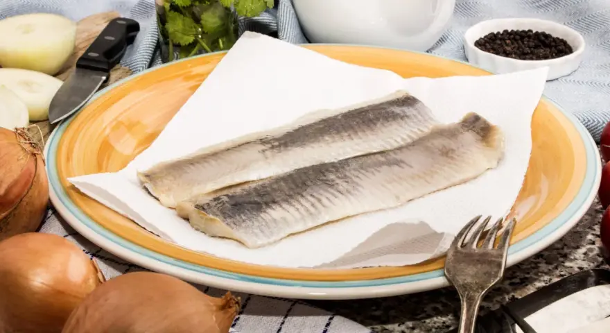 Zwei Matjesfilets auf Küchenpapier, bereit für eine gesunde Mahlzeit. Zwiebeln und Pfefferkörner deuten auf die Zubereitung hin.