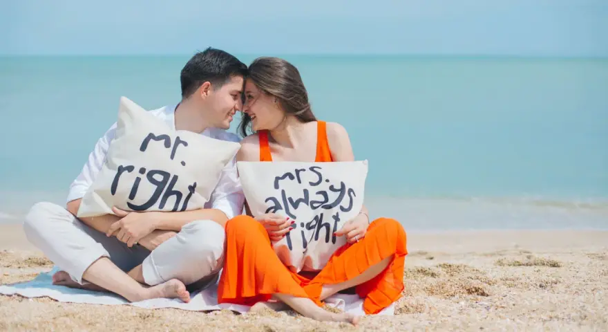 Para zakochanych na plaży z poduszkami "Mr. Right" i "Mrs. Always Right". Ich uśmiechy i bliskość symbolizują idealny **kompromis w związku**.