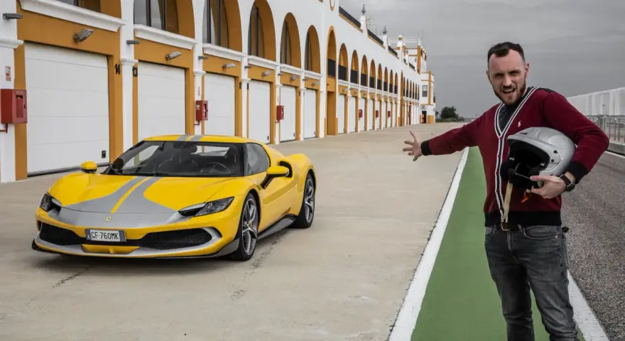 Żółte Ferrari 296 GTB z szarymi pasami na torze wyścigowym. Obok stoi mężczyzna z kaskiem, zapraszając do przejażdżki.