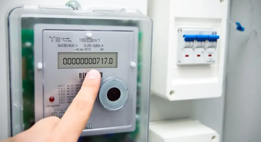 Palec wskazuje na cyfrowy licznik energii elektrycznej, pokazujący odczyt 00000000717.0. Widoczne są symbole na liczniku energii elektrycznej.