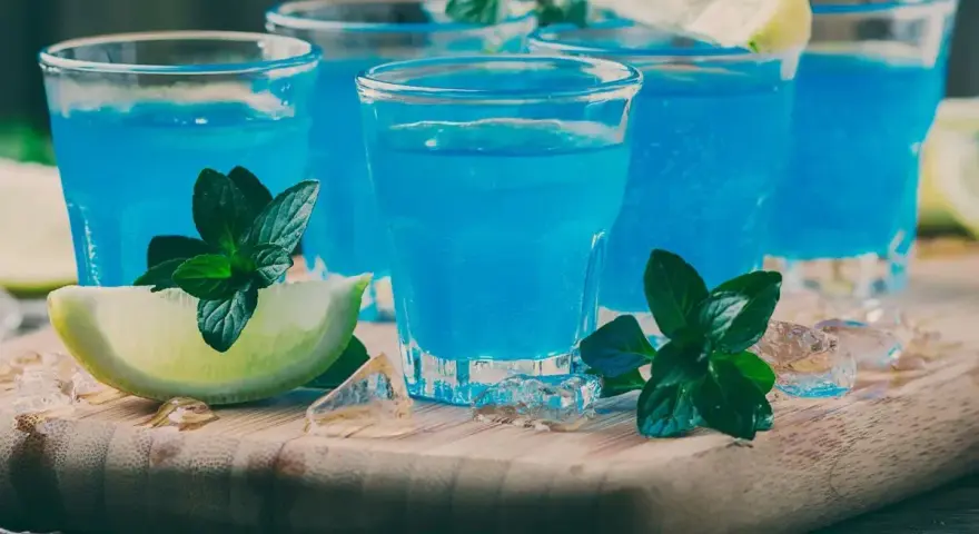 Kilka shotów z drinkiem blue curacao, ozdobionych limonką i miętą, na drewnianej desce z lodem.