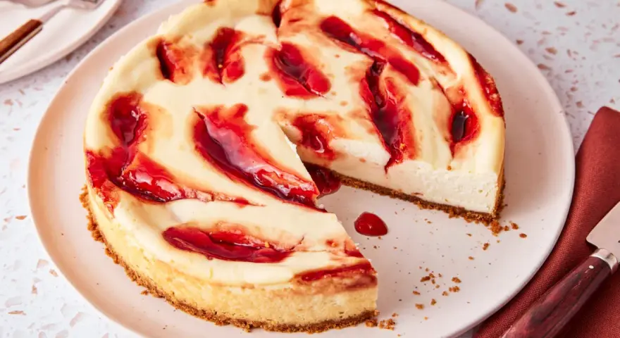 Ein saftiger american cheesecake mit keksboden, verziert mit roten Fruchtwirbeln. Ein Stück fehlt bereits.