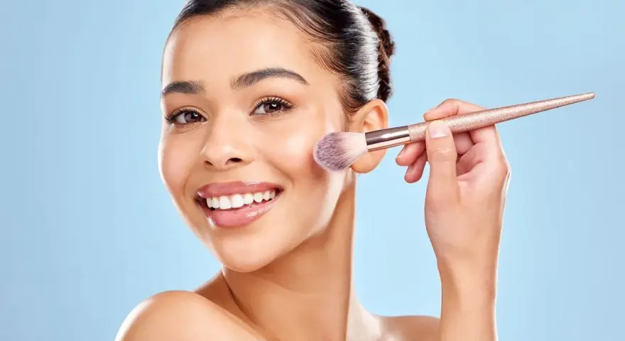 Une femme souriante utilise un pinceau pour appliquer du correcteur maquillage sur sa joue.